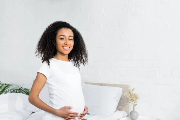 Mujer afroamericana feliz y embarazada sentada en el dormitorio - foto de stock