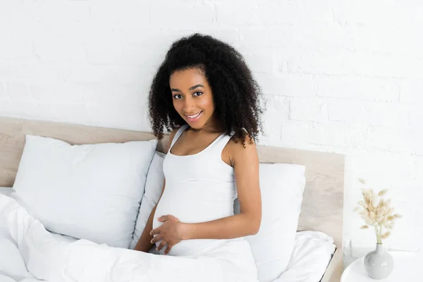 Embarazada africana americana mujer mirando a cámara, sentado en la cama en la habitación - foto de stock