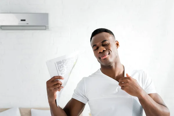 Homme afro-américain transpirant fermant les yeux, debout dans une pièce blanche avec température de chaleur, tenant le journal à la main — Photo de stock