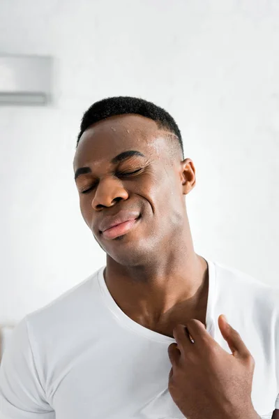 Hombre afroamericano agotado cerrando los ojos y de pie en la habitación con temperatura de calor - foto de stock