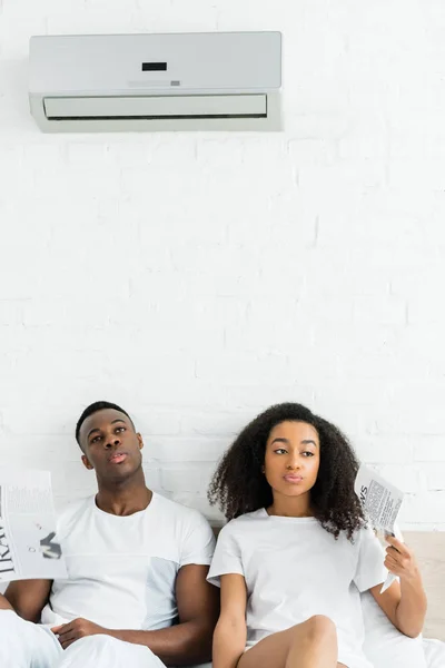 Müde afrikanisch-amerikanische Mann und Frau mit Zeitungen in den Händen auf dem Bett im Zimmer sitzen — Stockfoto