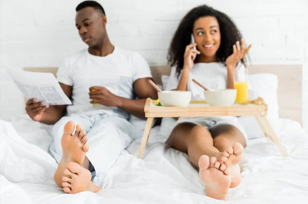 Selective focus of african american couple lying on bed, using smartphone and reading newspaper — Stock Photo