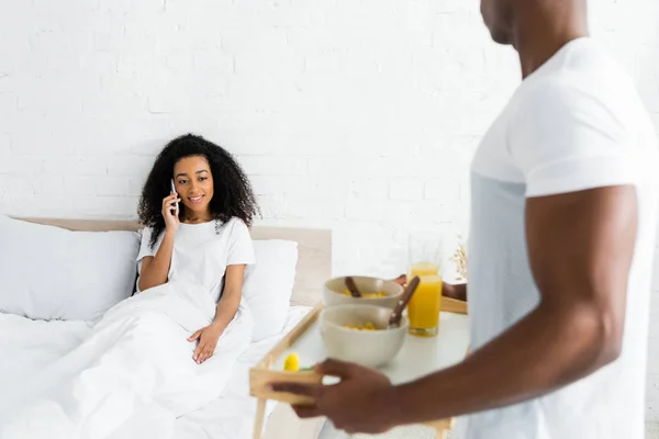 Enfoque selectivo de la mujer afroamericana hablando en el teléfono inteligente, mientras que el novio sostiene bandeja con desayuno - foto de stock