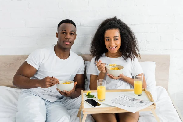 Alegre africano americano pareja comiendo desayuno en malo y mirando cámara - foto de stock