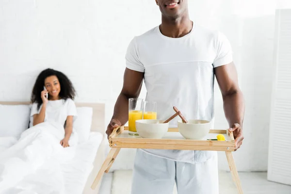 Vista selectiva del hombre afroamericano sosteniendo la bandeja del desayuno en las manos, con una novia sonriente en el fondo - foto de stock