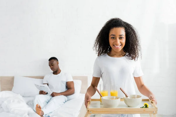 Fröhliche afrikanisch-amerikanische Frau hält Tablett in den Händen, mit Freund im Bett im Hintergrund — Stockfoto