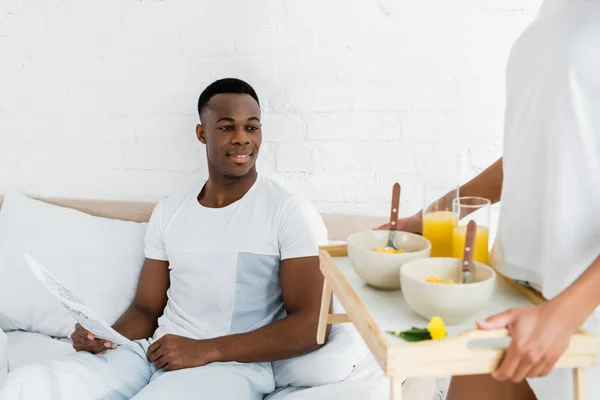 Happy african american man lying on white bed, and looking on tray in girlfriend hands — Stock Photo