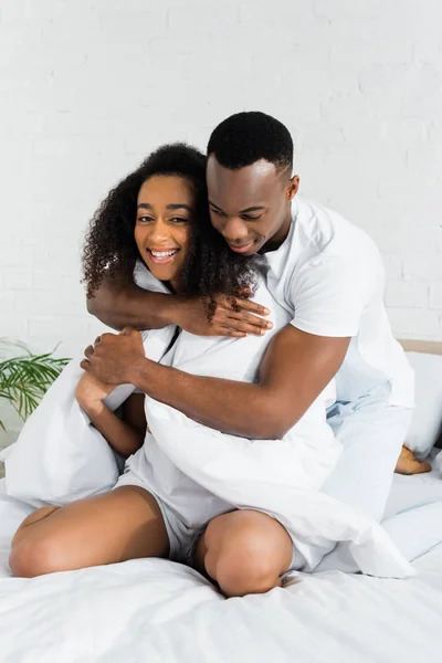 Afro-americano homem abraçando sua encantadora namorada, sentado na cama branca — Fotografia de Stock