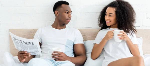 Vue de face du couple afro-américain couché au lit, se regardant, tenant tasse et journal — Photo de stock