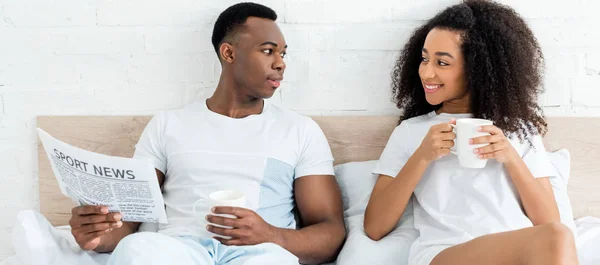 Vista frontal do casal afro-americano deitado na cama, olhando um para o outro, segurando copo e jornal — Fotografia de Stock