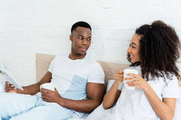 Feliz pareja afroamericana con periódico y taza acostados en la cama, mirándose el uno al otro - foto de stock