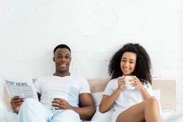 Heureux couple afro-américain regardant caméra, tenant des tasses blanches dans les mains — Photo de stock