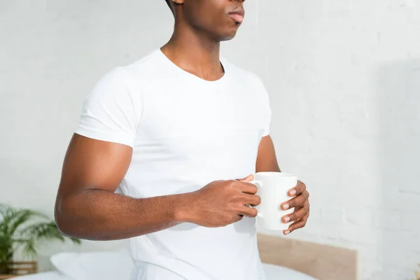 Vista recortada del hombre afroamericano sosteniendo la taza en las manos, de pie en la habitación blanca - foto de stock