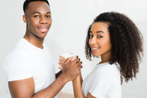 Heureux couple afro-américain regardant la caméra, tenant tasse avec boisson dans les mains — Photo de stock