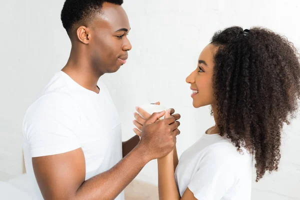 Vista lateral de la alegre pareja afroamericana mirándose, sosteniendo la taza con la bebida en las manos - foto de stock