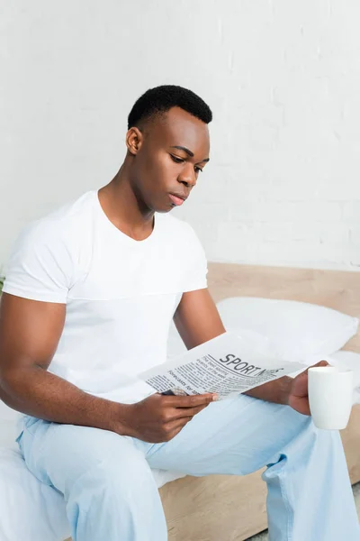 Hombre afroamericano inteligente sosteniendo la taza en la mano, leyendo periódico - foto de stock