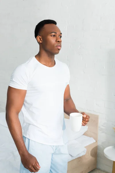 Hombre afroamericano serio de pie en la habitación blanca, sosteniendo taza con bebida en la mano - foto de stock