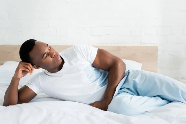 Hombre afroamericano tranquilo mirando hacia otro lado, acostado en la cama en habitación blanca - foto de stock