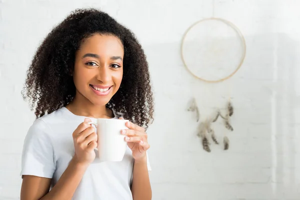 Alegre afroamericana mujer mirando a cámara con blanco taza en las manos - foto de stock