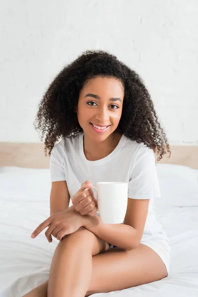 Glückliche afrikanisch-amerikanische Frau blickt in die Kamera und hält weiße Tasse in der Hand — Stockfoto