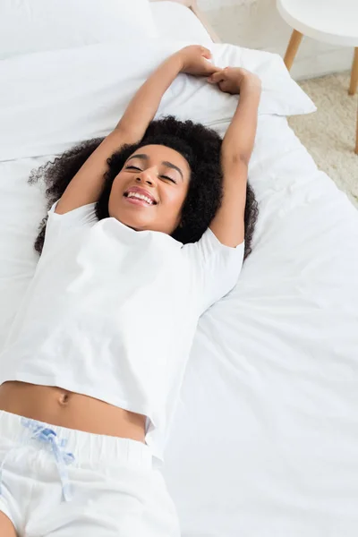 Vista ad alto angolo della donna afro-americana sorridente e sdraiata sul letto bianco — Foto stock