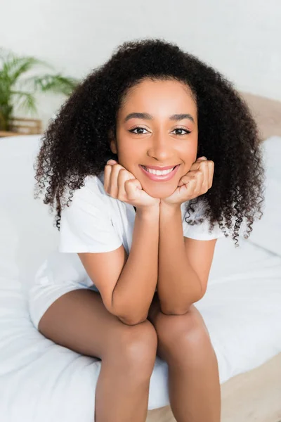 Vista de ángulo alto de la mujer afroamericana sonriendo, sentada en la cama en casa - foto de stock