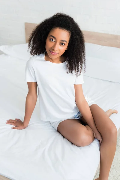 Vue grand angle de la femme afro-américaine regardant la caméra, assis sur un lit blanc à la maison — Photo de stock