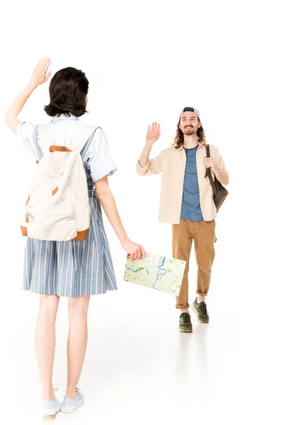 Visão de comprimento total da menina segurando mapa e mão acenando, enquanto o jovem caminhando e também acenando mão isolada no branco — Fotografia de Stock