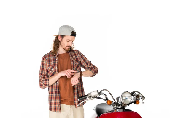 Молодий чоловік дивиться і вказує пальцем на годинник, стоячи біля червоного скутера ізольовано на білому — стокове фото