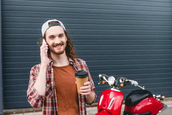 Молодой человек разговаривает по смартфону, держит бумажную чашку с кофе и стоит рядом с красным скутером — стоковое фото