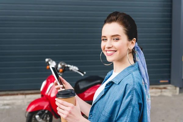 Belle fille debout près de scooter rouge, tenant tasse en papier avec café et smartphone — Photo de stock
