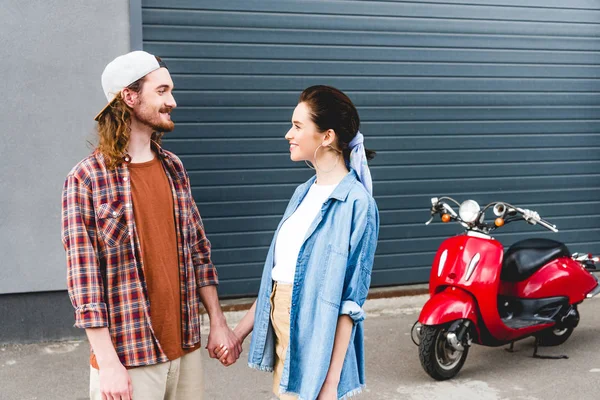 Joven hombre y chica mirándose el uno al otro y tomados de la mano con scooter rojo en backgroung - foto de stock