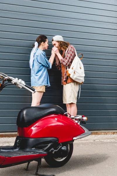 Hombre joven sosteniendo y besando las manos de la chica de pie cerca de la pared y scooter rojo — Stock Photo