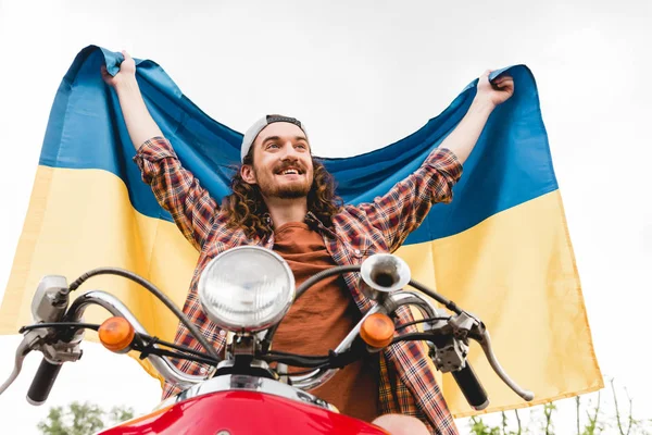 Vue à angle bas du jeune homme assis sur scooter rouge et tenant drapeau ukrainien — Photo de stock