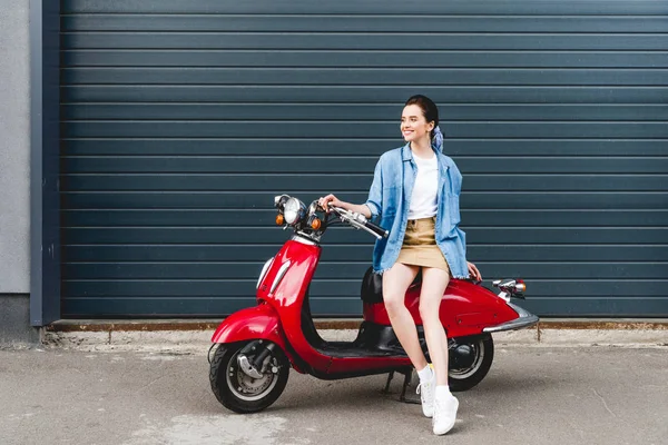 Вид в полный рост красивой девушки, сидящей на красном скутере и отводящей взгляд — стоковое фото