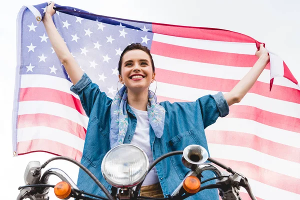 Low angle view of beautiful girl sitting on scooter, holding American flag and smiling on sky background — Stock Photo