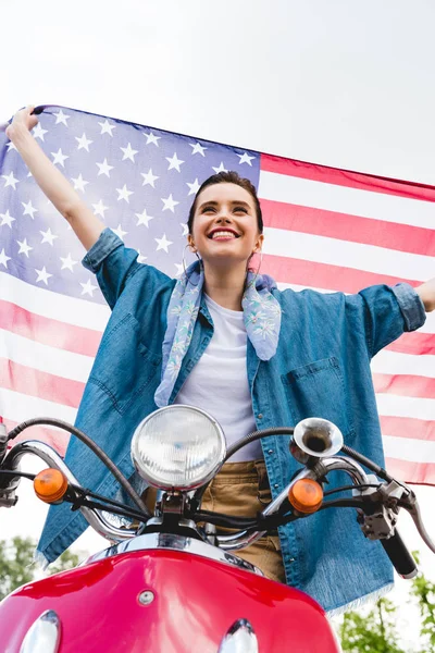 Niedrigwinkel-Ansicht des schönen Mädchens, das auf rotem Roller steht, amerikanische Flagge hält und auf Himmelhintergrund lächelt — Stockfoto