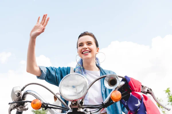 Low angle view of beautiful girl sitting on scooter , smiling and waving hand — Stock Photo
