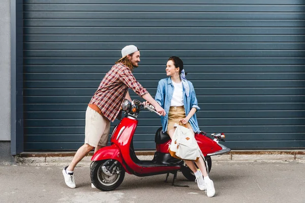 Вид в полный рост молодой пары, которая смотрит друг на друга и улыбается, сидя на красном скутере — стоковое фото