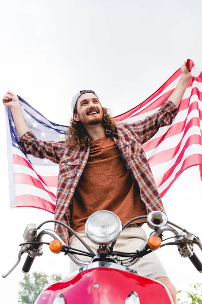 Low angle view of young man standing on red scooter, smiling and holding American flag — Stock Photo