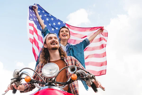 Low angle view of beautiful girl riding with young man on red scooter and holding American flag — Stock Photo