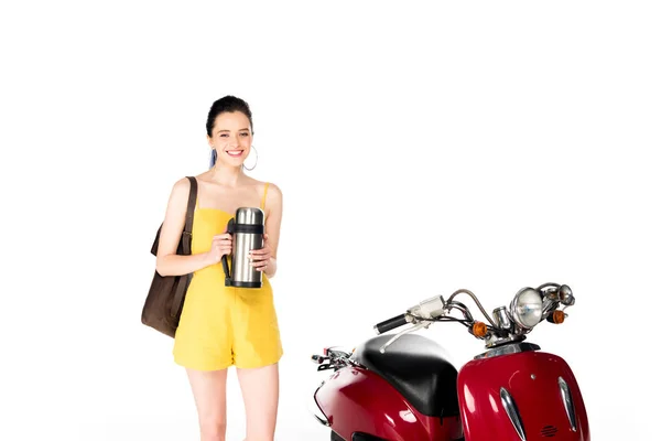 Menina bonita em roupas amarelas segurando mochila e garrafa térmica, olhando para a câmera e de pé perto scooter vermelho isolado no branco — Fotografia de Stock