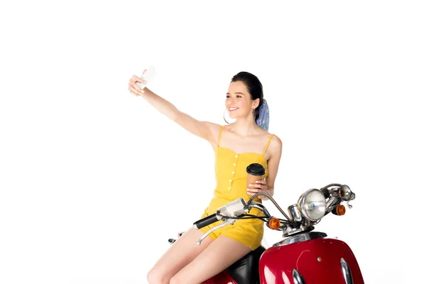 Beautiful girl in yellow clothes holding paper cup, taking selfie and sitting on red scooter isolated on white — Stock Photo