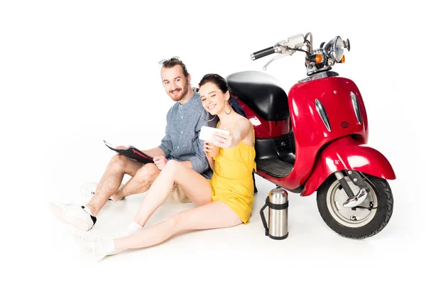 Joven hombre y chica sentado en el suelo cerca de scooter rojo y tomar selfie aislado en blanco - foto de stock