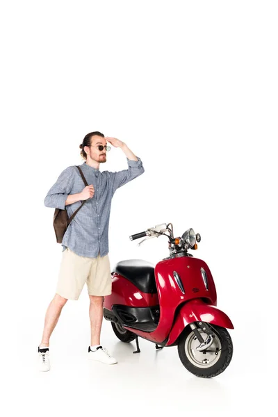 Vue pleine longueur de beau jeune homme élégant avec sac à dos debout près de scooter rouge et regardant loin isolé sur blanc — Photo de stock