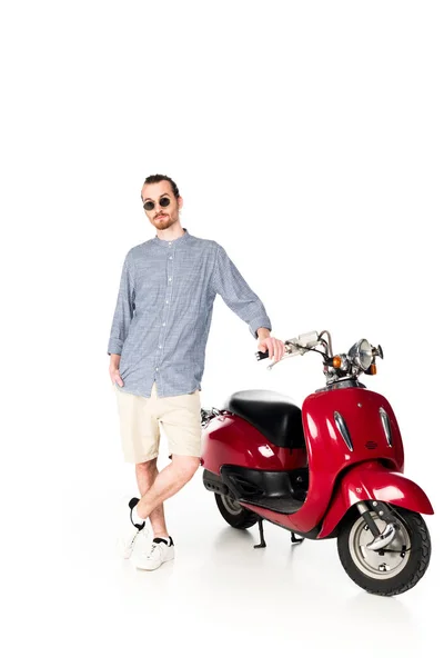 Ganzkörperansicht des hübschen jungen Mannes, der in der Nähe eines roten Motorrollers steht und die Kamera isoliert auf weißem Grund betrachtet — Stockfoto