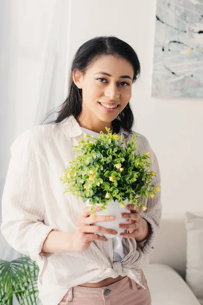 Jolie femme latine tenant pot de fleurs avec plante verte tout en souriant à la caméra — Photo de stock