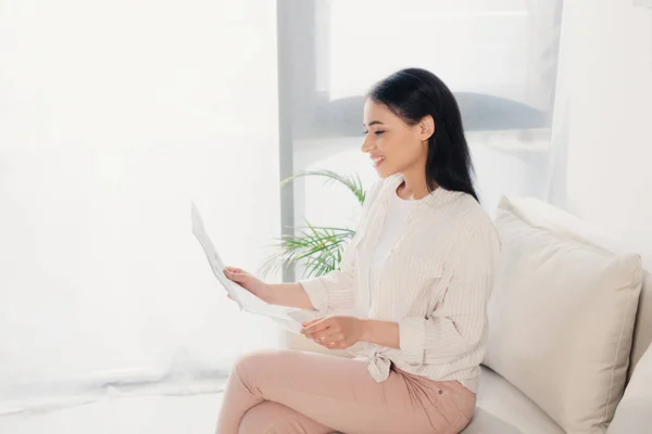 Alegre mujer latina leyendo el periódico mientras está sentada en el sofá en casa - foto de stock