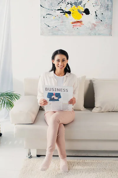 Beautiful latin woman holding business newspaper while sitting on couch and smiling at camera — Stock Photo