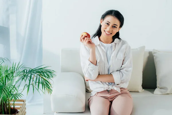 Belle femme latine tenant pomme mûre tout en étant assis sur le canapé et souriant à la caméra — Photo de stock